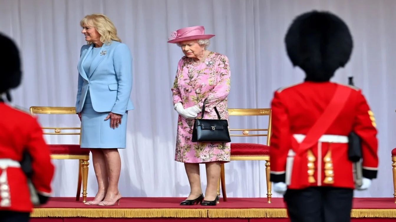 Jill Biden in blue suit and Elizabeth II in pink floral dress stand on stage with guards.