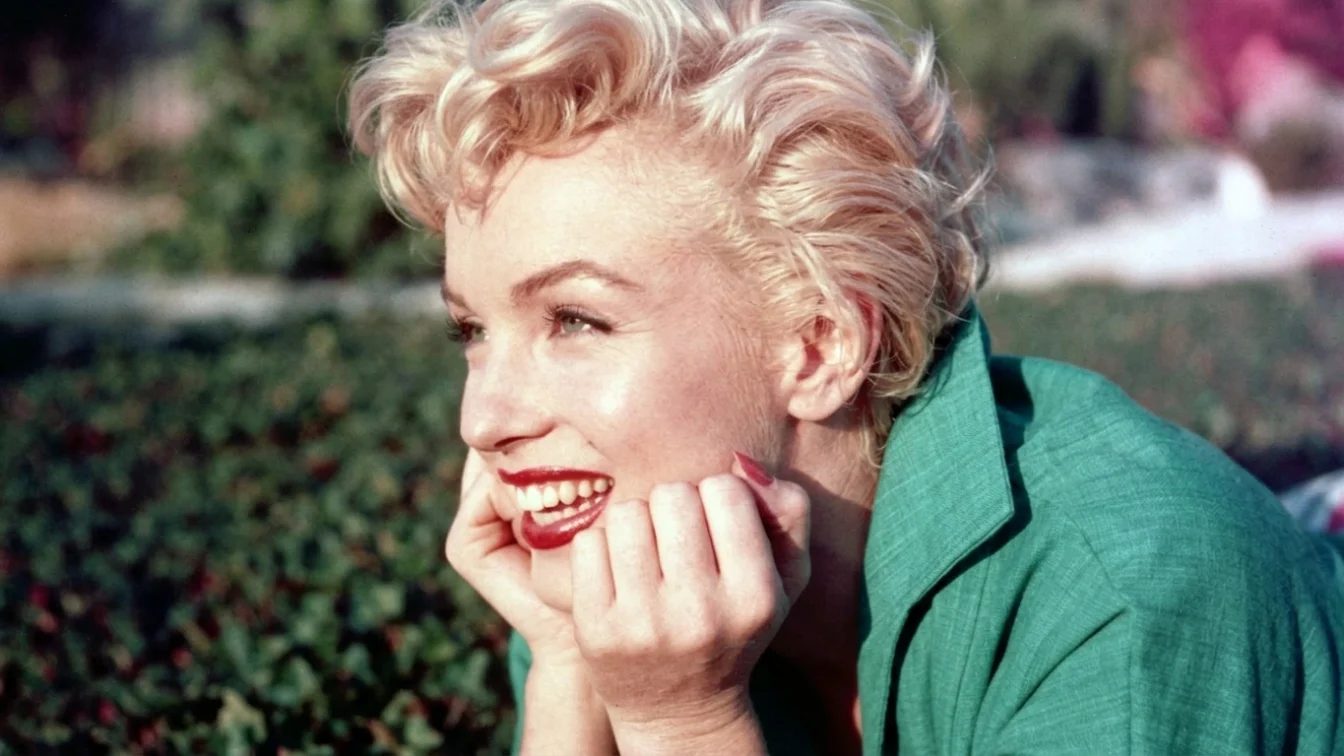Marilyn Monroe poses in the grass in a green shirt.