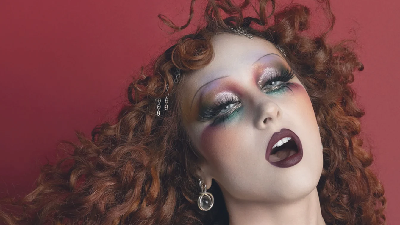 Chappell Roan appears in a MAC Viva Glam ad wearing a flowing costume and holding a flag. Her hair is styled in curls...
