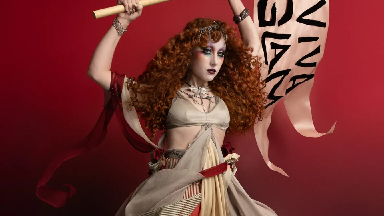 Chappell Roan appears in a MAC Viva Glam ad wearing a flowing costume and holding a flag. Her hair is styled in curls...