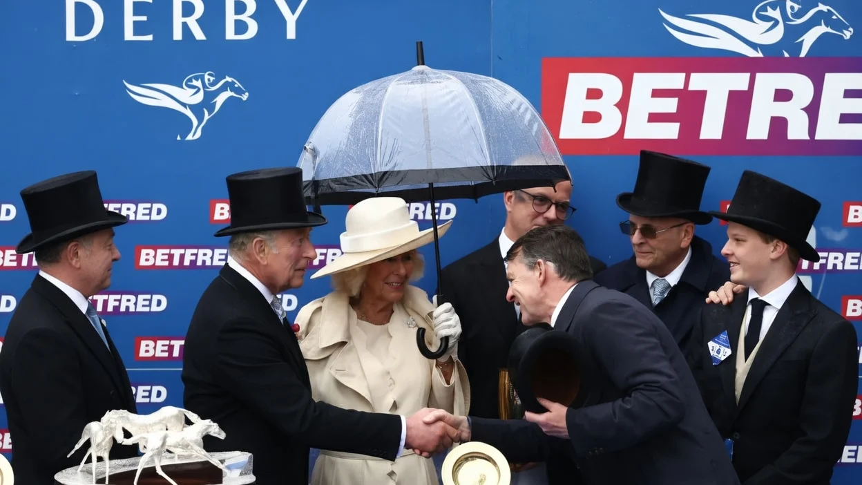 King Charles shaking hands with a horse trainer while standing with Queen Camilla