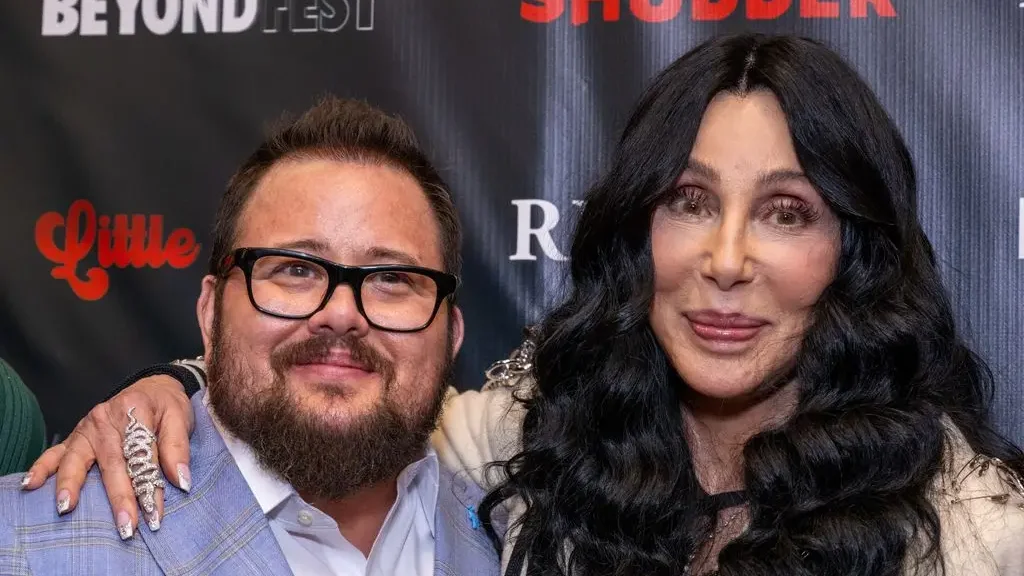Chaz Bono in a blue suit and white shirt, standing next to his mother Cher who holds an arm around her son.
